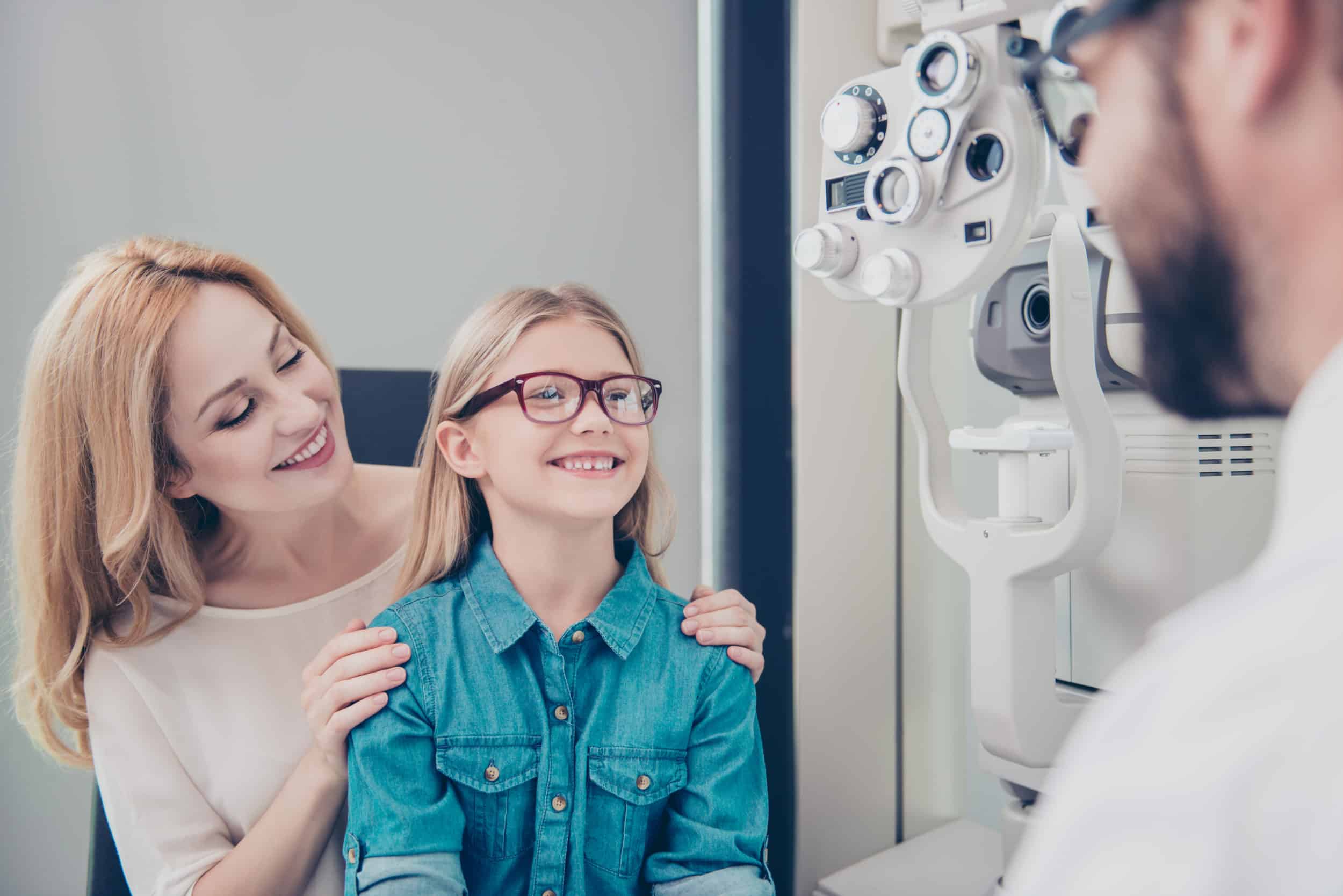 Healthcare, people, eyesight and vision concept. Small cute blond girl is trying on glasses, mom and doc are smiling, she is happy